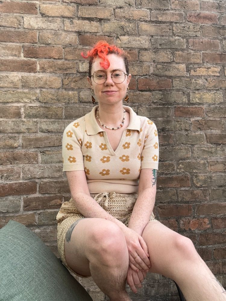 Lydia is a white person with brown hair and orange dyed bangs. She wears a cream polo with yellow daisies and tan marled crochet shorts.