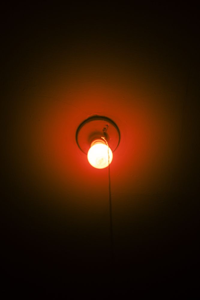 A color photo of an exposed lightbulb with a pull string that is on and has a red hue. 

Malden, MA. 2025.
