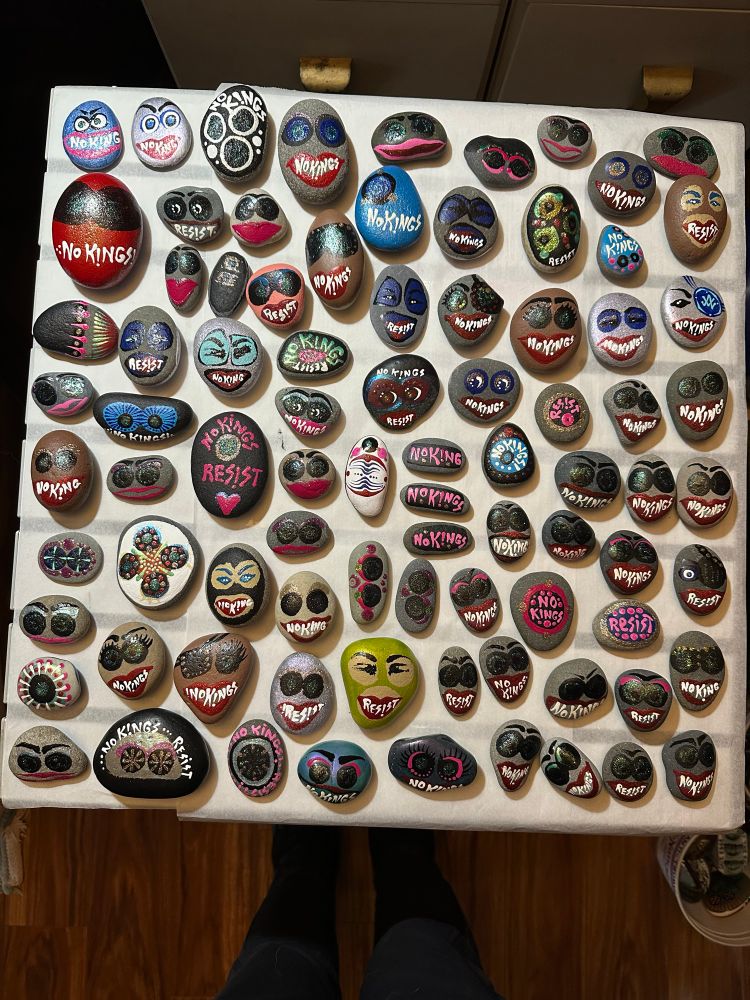 Painted rocks that have faces in balaclavas or red lipstick and white teeth spelling out: No Kings or Resist! The rocks are left laying around in an attempt to penetrate social media silos in a MAGA Trump town. 