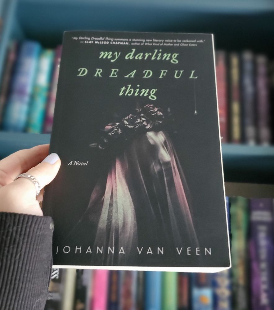 A left hand holds My Darling Dreadful Thing by Johanna Van Veen. It's a black cover with a veiled person wearing a black flower crown.