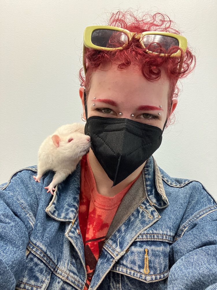 Red-haired person with mask on, an albino rat posed on their shoulder to the left. He is leaning towards the face, sniffing the mask.