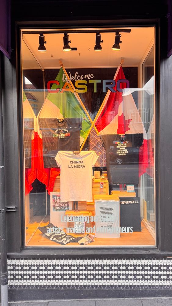 Store window with Welcome Castro on the glass, and a display with Mexican and Canadian flags behind several message t-shirts with the sayings, “chinga la migra,” “stonewall,” “but it was assigned Gulf of Mexico at birth,” and “Diversity hurts no one, you stupid orange f*ck.”