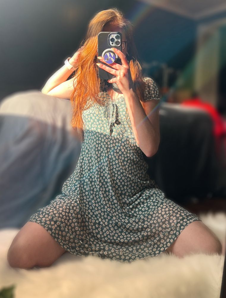 I’m kneeling on a fuzzy white rug in front of a mirror, wearing a short sundress dress. The photo’s taken in soft, warm light with a rainbow lens flare from the light hitting just right. My legs are spread slightly as I pose with one hand in my hair and the other holding my phone, partially covering my face. 🖤🧡