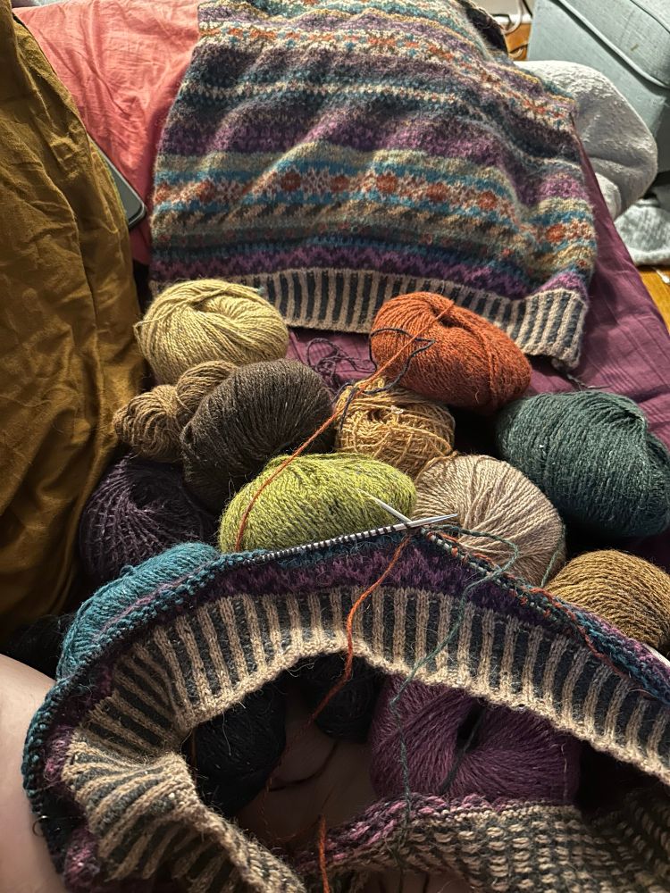 Twelve different colours balls of yarn spread out with a work in progress knitted cardigan project with just the bottom hem and first few colourwork rows complete. In the background a finished back of the same cardigan pattern representing a past attempt to knit this. 