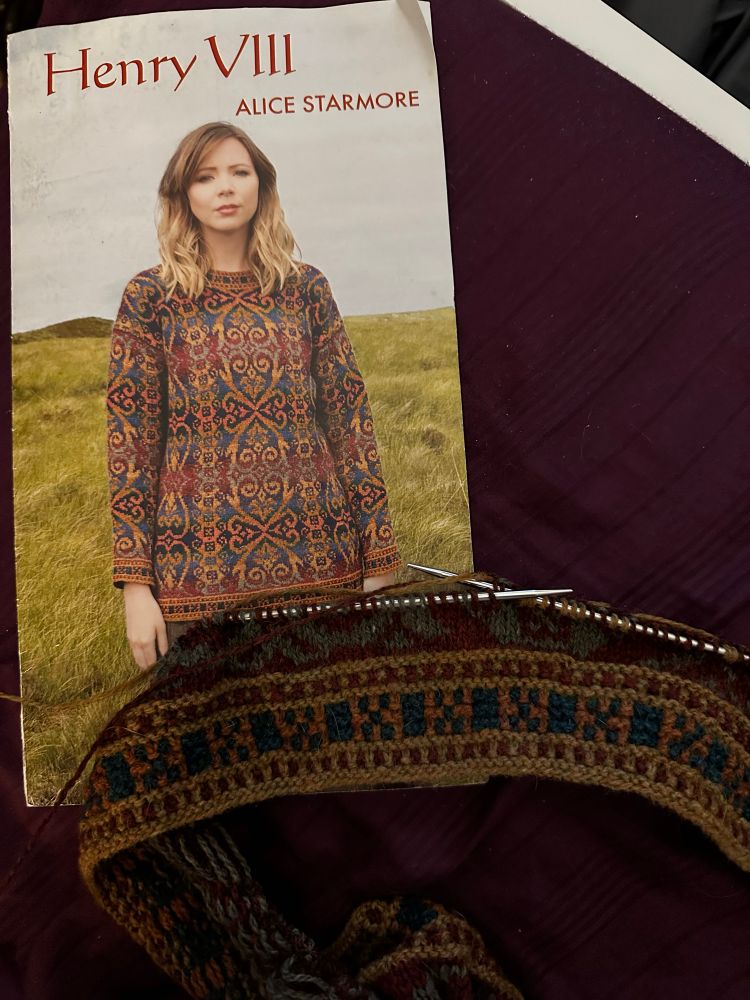 A pattern booklet showing a tapestry like sweater in rich reds, gold, blues, and greens and a work in progress knitting project on metal needles 