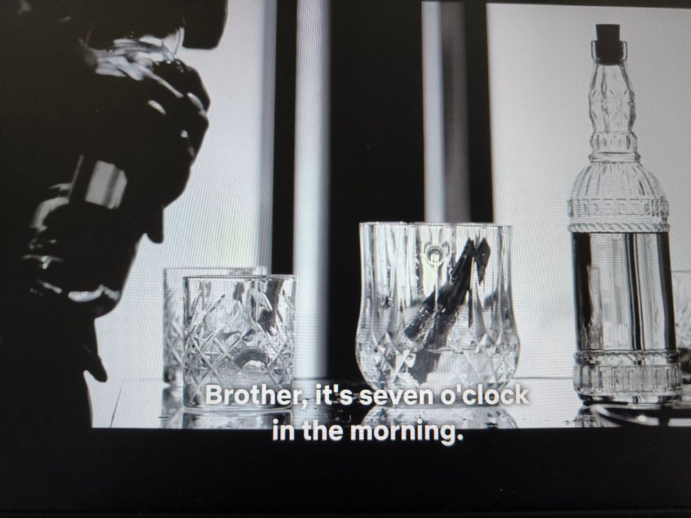 Photo de l'écran de mon pc sur lequel je regarde Animal, un film indien. C'est un gros plan de verres en crystal et bouteilles d'alcool sur une table. L'image est en noir et blanc. Une main débouche une bouteille. Les sous-titres affichent, en anglais: "Frère, il est 7 heures du matin."