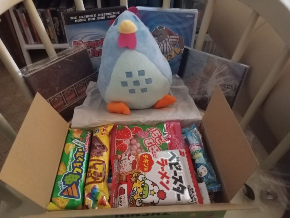 A whole load of nice gifts! Beat the intro and bullseye dvd games, YMO's kyoretsu na rhythm and Oingo Boingo's Boingo on cd, a blue chicken plush and a box of Japanese snacks. Next to the box are a couple of small pins which are hard to see, but there's a cat one and a starfleet one with a hello kitty one not pictured