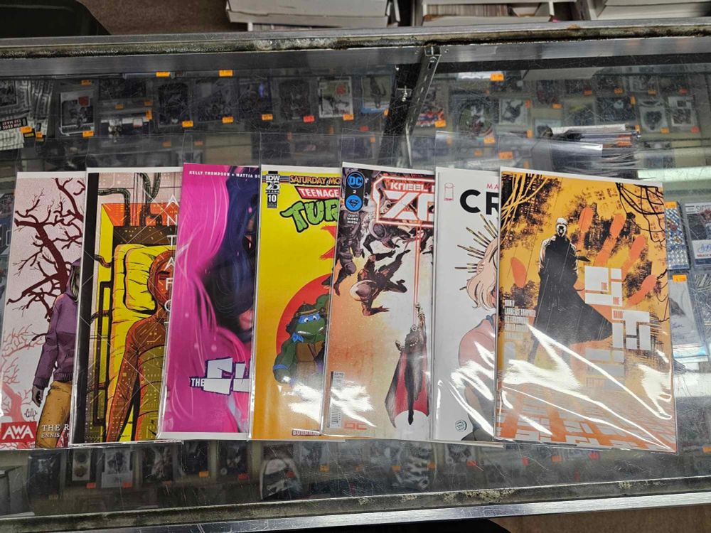 A sprawl of comics on a glass top counter. The comics are 
Ribbon queen
Antarctica
Saturday morning TMNT
Kneel before zone
Crave
The one hand