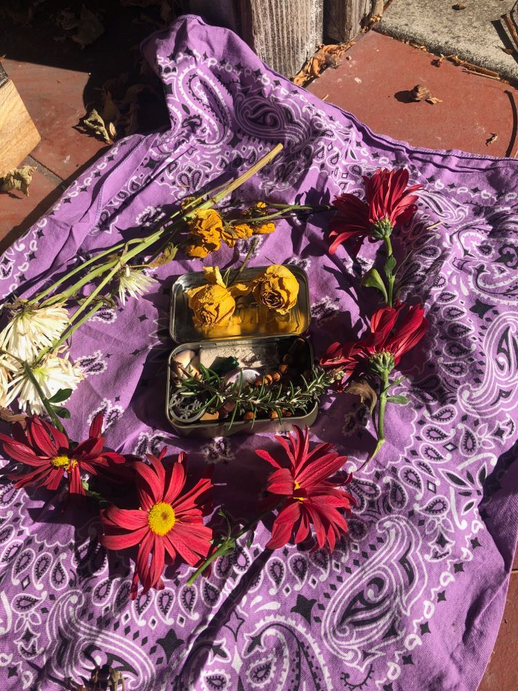 Assembling my grief bundle atop a lavender hankie. I'm not going to explain the meaning behind the items visible, but there're some wooden prayer beads, a phial of ashes, some rosemary, some seashells, and some old jewelry visible. The Altoids tin they're being assembled in is ringed with flowers from my altar.
