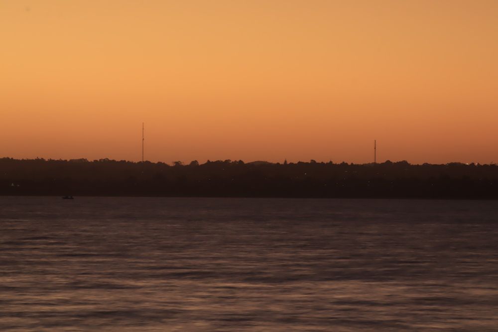 Zoom photo of Te-Atatu peninsula with two radio masts at left and right of centre. Peninsula is in silhouette across the middle of photo. orange and yellow sky is in upper half, while dark ocean with streaks of faint yellow is in lower half.