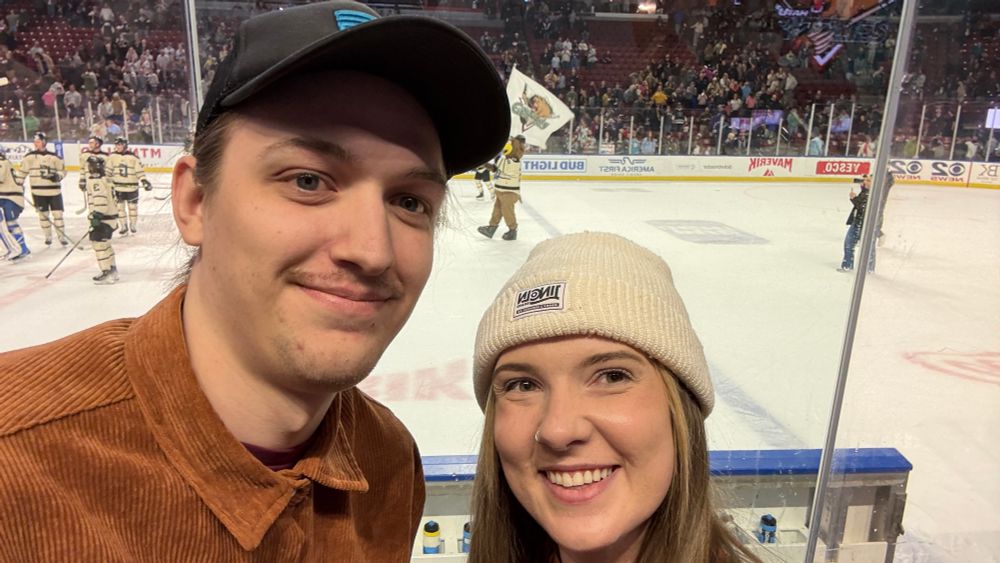 a blonde haired woman and brunette man standing in front of an ice rink, behind the bench for the home team, the Utah Grizzlies. the mascot, Grizzbee, a grizzly bear, is out on the ice with the team members celebrating their win against the Kansas City Mavericks. Grizzbee is waving the Utah Grizzlies flag. 
