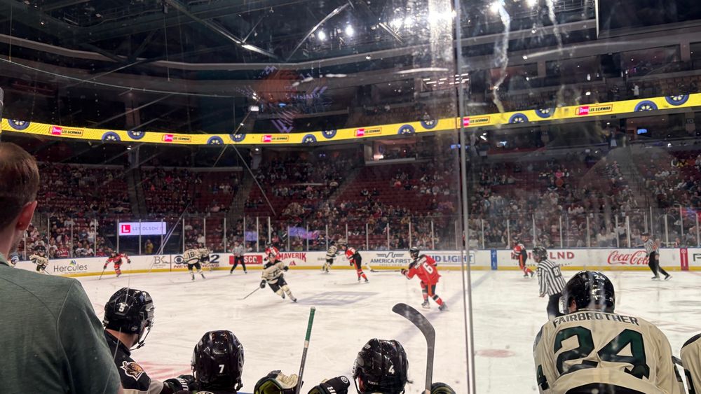 a photo of an ice arena taken from directly behind the home team bench. you can see part of one of the coaches on the left hand side of the screen, 4 players on the bench, and all the players on the ice. they are mid-play, and i am unsure of who has possession of the puck as it is unclear from the photo. 