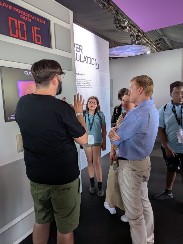 Researcher Dr Will Roper is on the left, talking to Astronaut Tim Peake featured on the right. They are in the Euclid exhibition, seen in the background. In particular they are looking at an interactive screen of a Universe simulator called Swift.