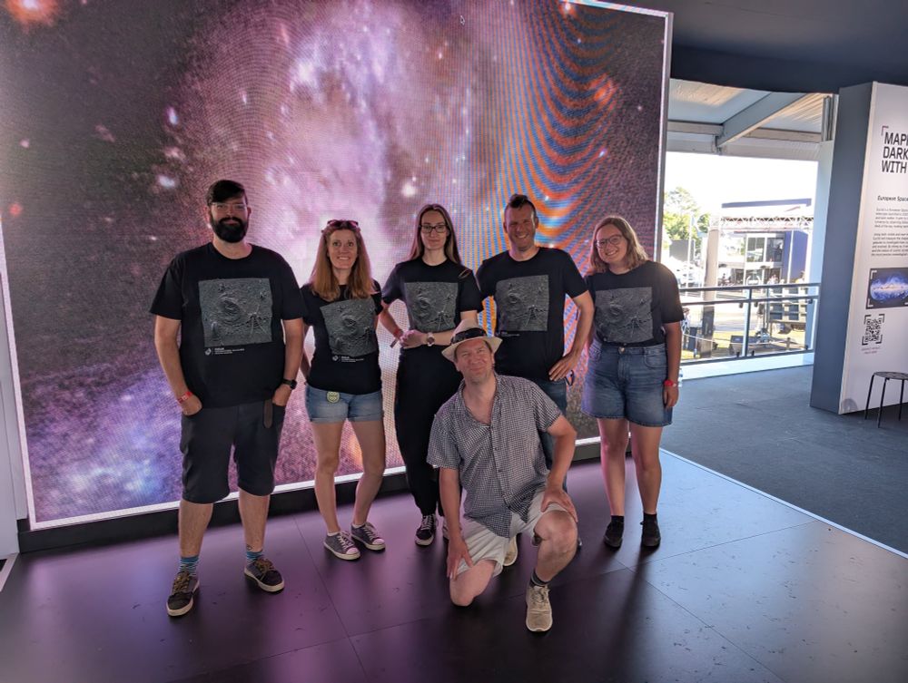 Some of the Euclid team at the Euclid exhibit. Five people are standing together smiling for the camera. One person is knelt in front of them, also smiling for the camera. Behind them in the background is a spiral galaxy, one of Euclid’s images displayed on a huge screen. 