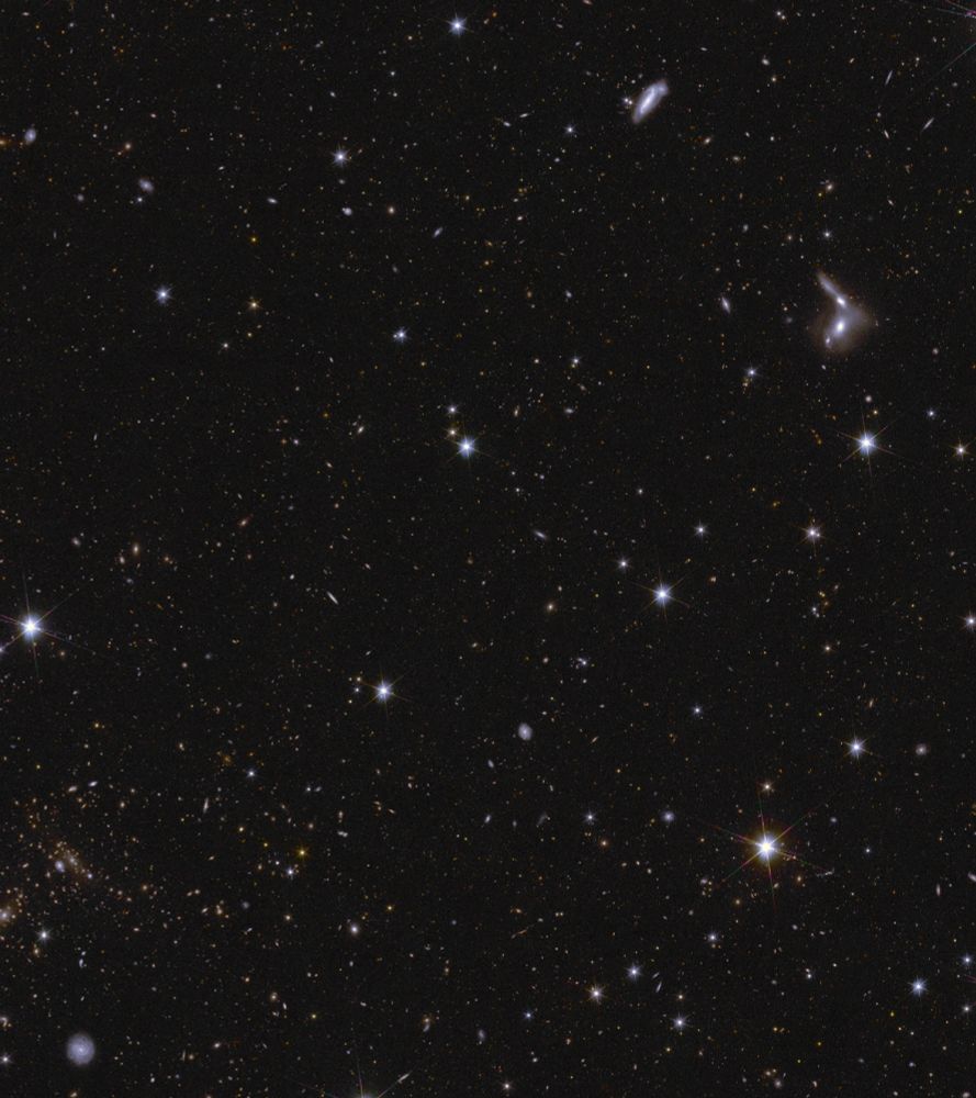 RIGHT - Image description: A sea of stars and galaxies sparkle against a black background. Several galaxies can be identified by their elongated shape and/or spiral arms. Some galaxies are seen edge-on while one prominent spiral galaxy at the bottom centre is seen face-on. At the far right, between the middle and top of the image, are some interacting galaxies. Galaxy clusters are also seen, in particular near bottom centre, where features smeared into arcs represent gravitational lensing. The brightest stars in the image have diffraction spikes.]