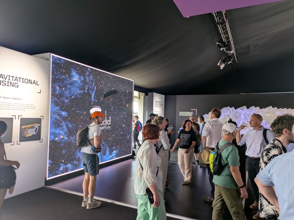 A wide shot of part of the Euclid exhibition at the Goodwood Festival of Speed. In the foreground are lots of people wandering around the exhibit. On the left is a huge screen displaying a Euclid image. The exhibition is held in a large black tent, which drapes from the top of the image. 