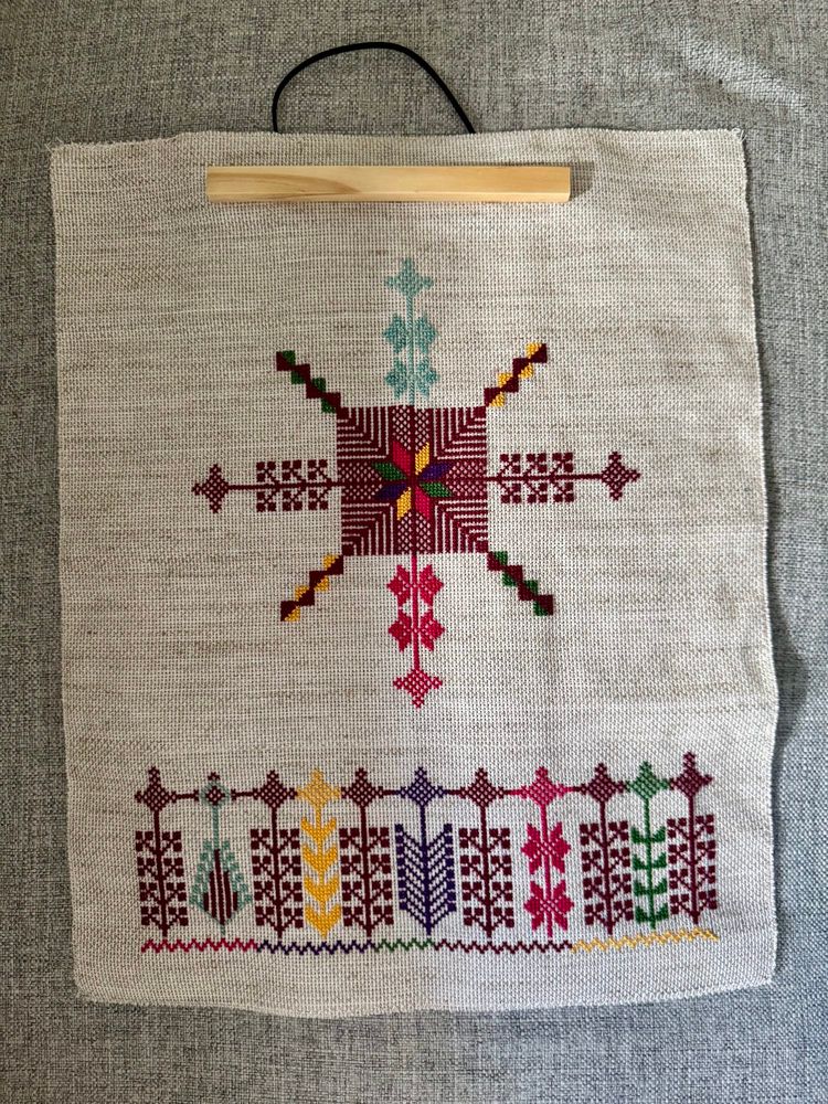Tatreez embroidery (cross-stitch style from the Levant) on a natural aida cloth. The central motif is a feathered moon, mainly stitched in dark red with pink, teal, yellow and green accents. Below the central motif is a row of various tree and plant motifs that form a wall. 