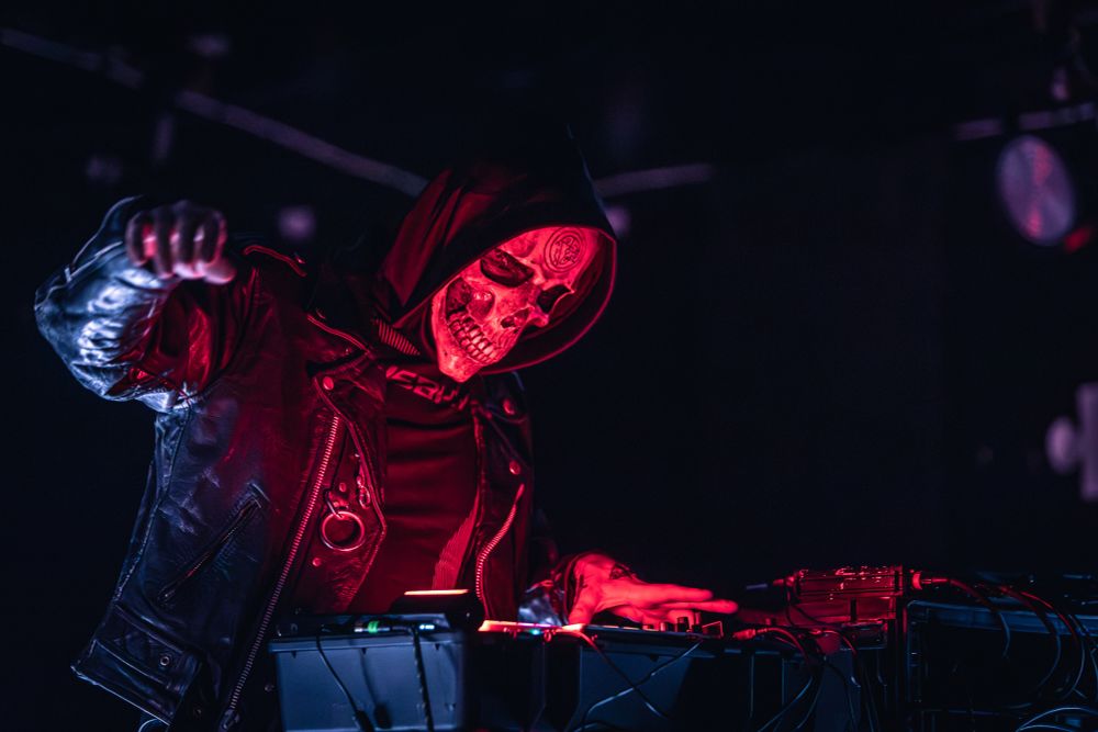 One member of musical act Gost performing at X-Ray Arcade in Milwaukee, WI; one arm is outstretched as he operates electronic music equipment. The room is dimly lit with dramatic red lighting accents.