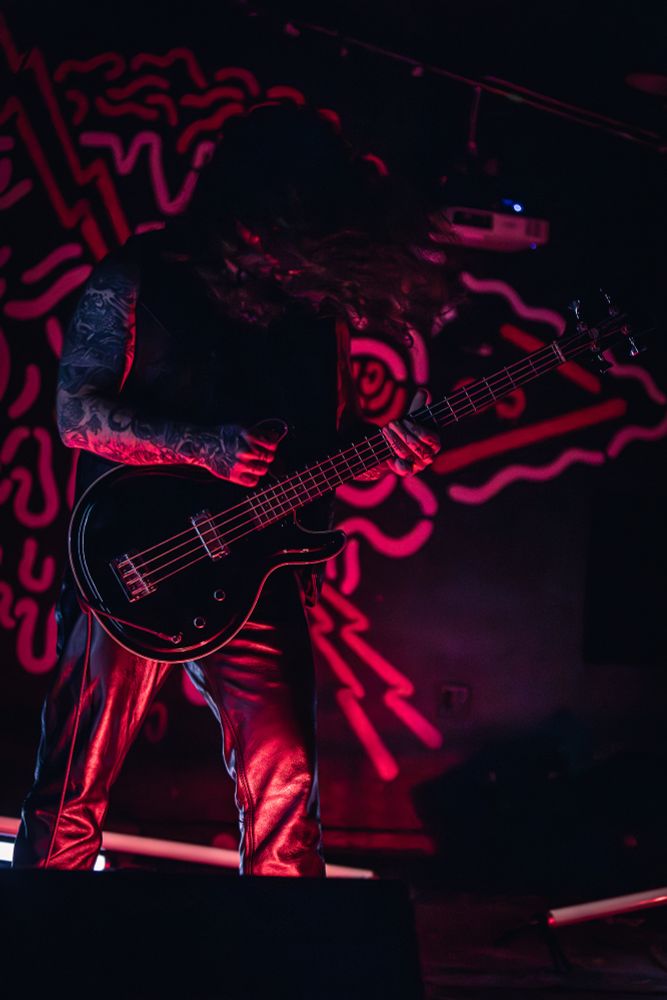 The second member of musical act Gost performing at X-Ray Arcade in Milwaukee, WI; he is mid-strum on a bass guitar while his long hair is in motion. The room is dimly lit with dramatic red lighting accents.