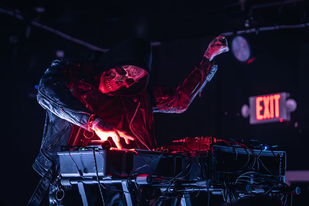 One member of musical act Gost performing at X-Ray Arcade in Milwaukee, WI; his clenched fist is raised menacingly as he operates electronic music equipment. The room is dimly lit with dramatic red lighting accents.