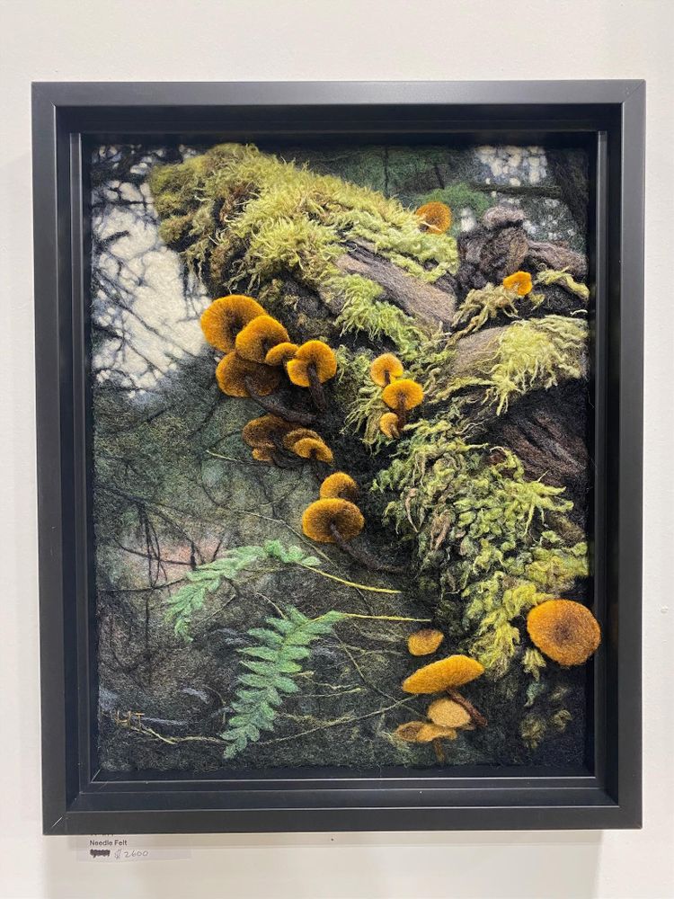 needle felted, photorealistic wall sculpture of a mossy log covered with orange mushrooms, with trees in the background. Based on a photo taken in North Vancouver along Seymour River on Fisherman's Trail