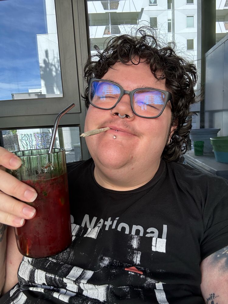 Theo wears black glasses and a black The National T-shirt, with a joint in their mouth and a pink drink in a green glass in their hand. they are smiling with their eyes closed
