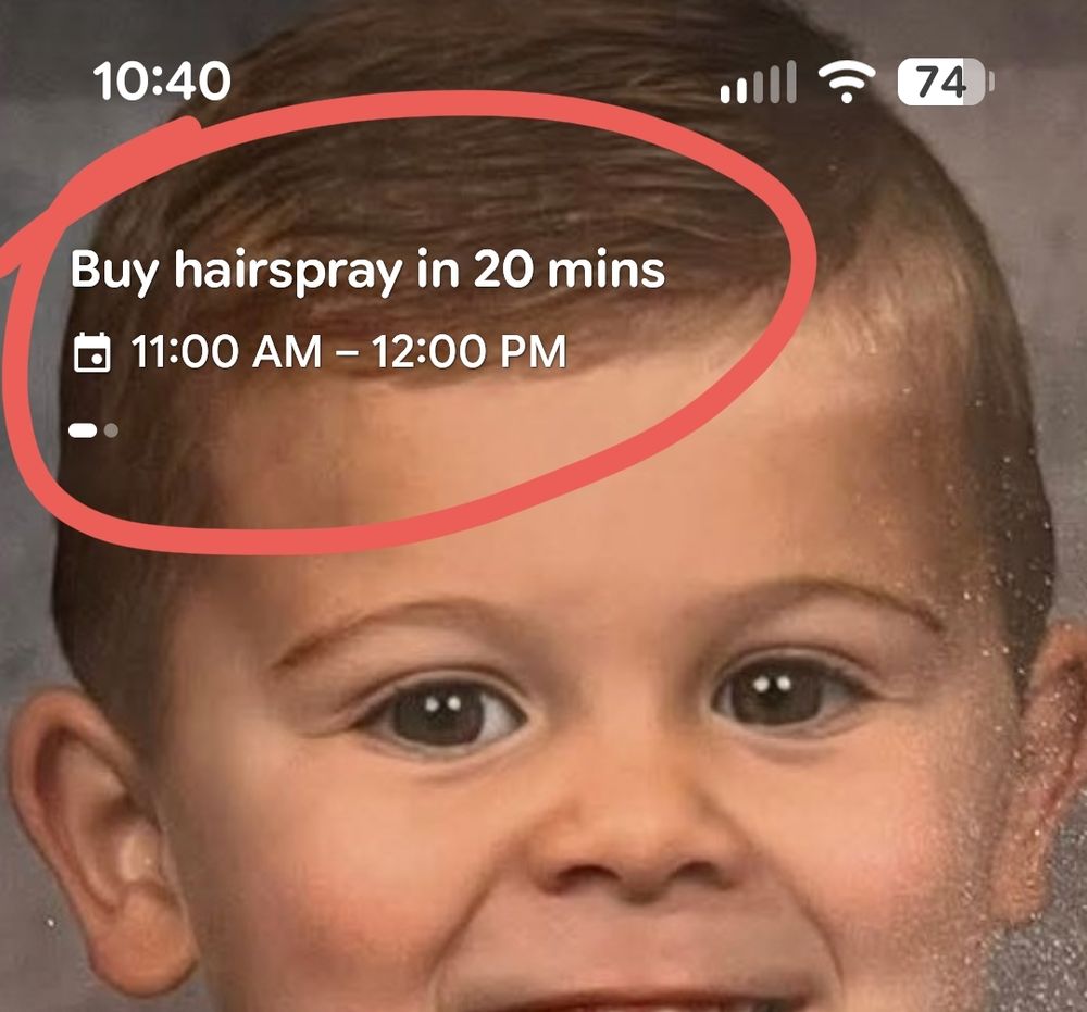 Screenshot of my phone with the face of a smiling toddler as wallpaper and on his brown hair, white letters say "Buy hairspray in 20 mins"