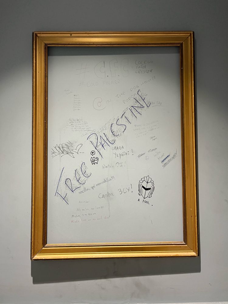 A golden frame surrounding a blank space on the wall in a public bathroom where someone bas scribbled Free Palestine in a prominent diagonal line.