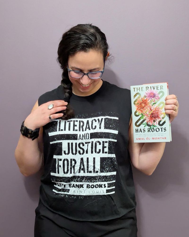 Photo of me holding up a copy of THE RIVER HAS ROOTS to the right while looking down at my shirt and smiling. The shirt reads LITERACY AND JUSTICE FOR ALL in black, block, stencil-style black letters encased in white against the black of the shirt. Beneath that it says “Left Bank Books, St Louis.” It was once a t-shirt but is now a tank. My hair’s in a side braid and I’m wearing an oval moonstone ring and a black leather cuff. On the hand not holding the book.