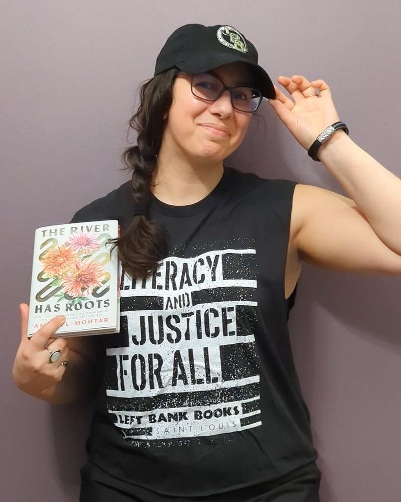 Same outfit as the previous photo but the book has shifted to the left side of the photo as I’ve added an amazing black baseball cap with Left Bank Books’ logo (LBB) in a circle, and have raised my free hand to touch its brim while looking amused and pleased with myself.