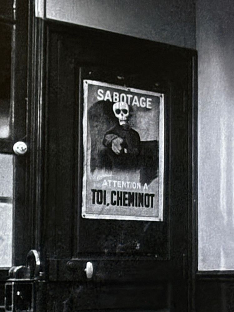On a black door, a poster featuring a death’s head clad in black and pointing at the viewer. Above it is the word SABOTAGE and below it, in French “Attention à Toi, Cheminot,” meaning “Watch Yourself, Railwayman.”