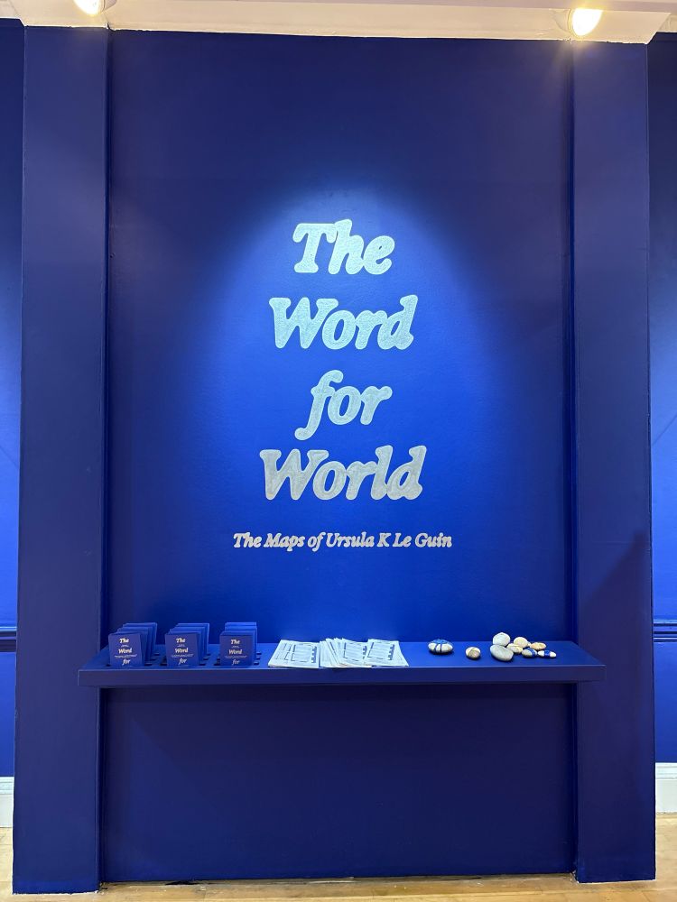 A dark blue wall on which the words THE WORD FOR WORLD are spotlit in white, subtitled “The Maps of Ursula K. Le Guin.” Beneath them is a shelf on which are maps for navigating the exhibit as well as maps illustrating its contents: to the right is a pile of stones inscribed with designs or covered in knotwork, to show how maps are talismans in Le Guin’s novel ALWAYS COMING HOME.