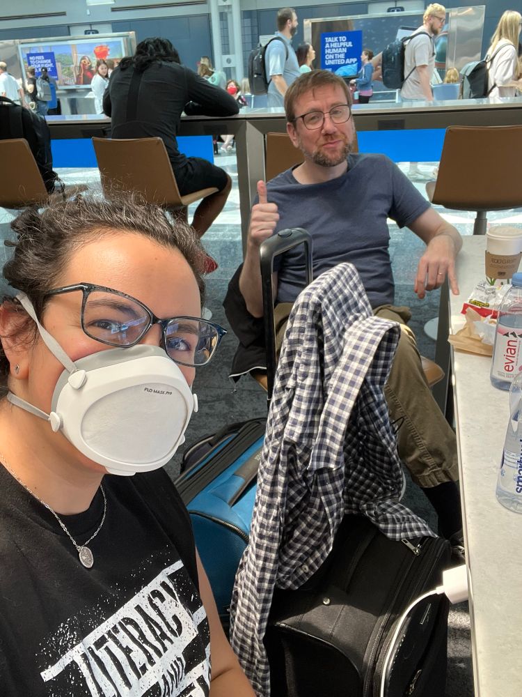 Airport selfie including my husband. I’m wearing a Flo mask, glasses, and a black shirt that reads “Literacy and Justice for All,” while my husband, a white man with short light hair and a salt and ginger beard, is wearing a dark blue short and giving an ironic thumbs up. He’s unintentionally but hilariously juxtaposed with a blue sign in the background that reads “Talk to a helpful human on the United App”