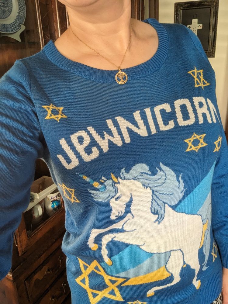 me wearing a blue holiday sweater featuring Stars of David and a unicorn superimposed on a cyan, blue, and yellow rainbow, with the word "jewnicorn" across the top