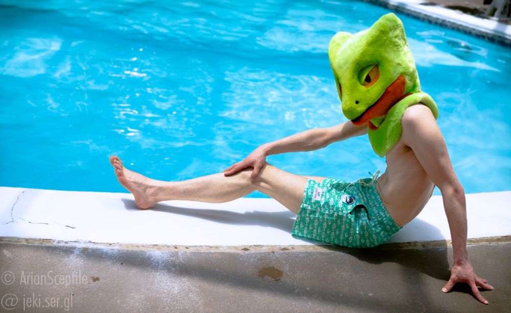 A sceptile fursuiter lounging by the pool in teal swimming trunks.