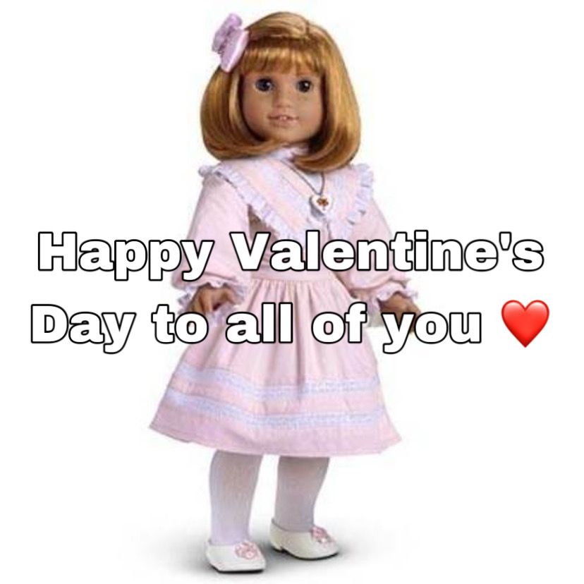 A White doll with blue eyes, freckles, and shoulder length red hair with a pink bow clip wearing a light pink dress with white lace trim and white ruffles, white tights, a silver chain with a white heart pendant, and white ballet flats with pink flower shaped bows on the toes stands in front of a plain white background with her body facing slightly to the right and her head facing the center. The image text says "Happy Valentine's Day to all of you" with a red heart emoticon at the end of the sentence
