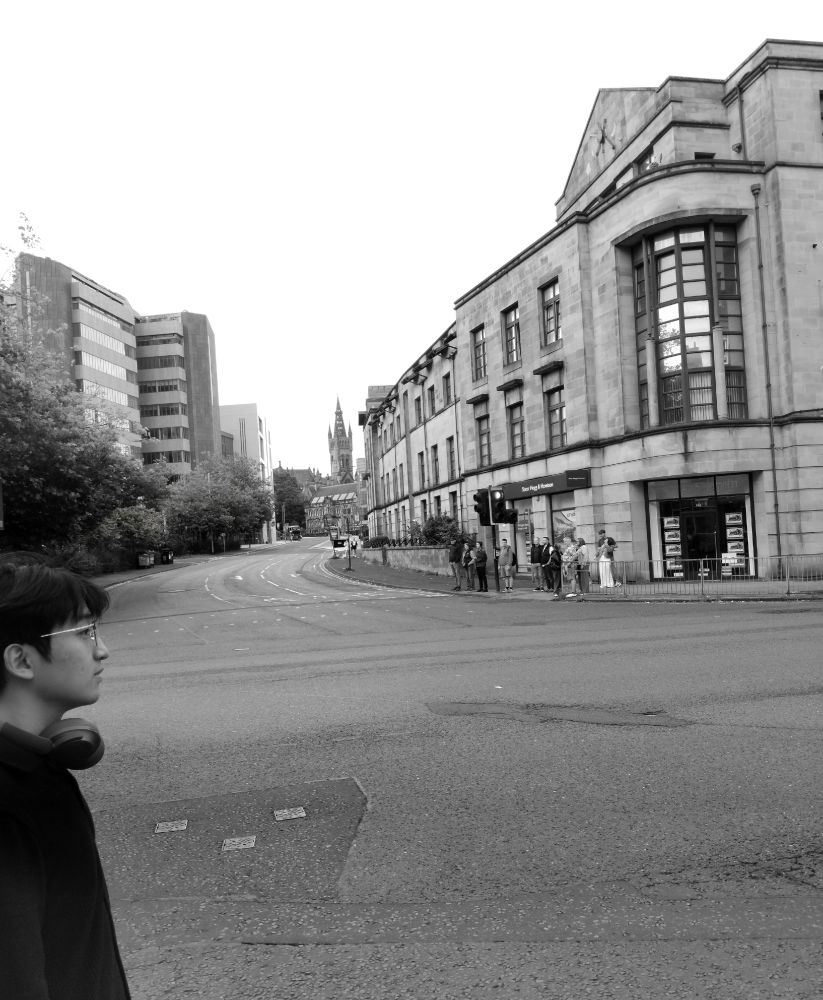 A monochrome photograph showing a bespectacled young man waiting to cross the street at a city intersection. He is in profile on the left of the picture, with headphones around his neck, and we cannot see the back of his head or anything below his chest. Diagonally opposite him, a group of people are also waiting to cross.