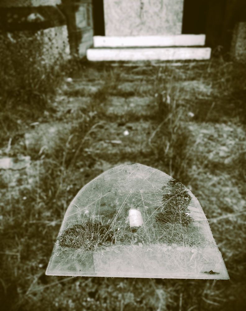 A monochrome photograph showing a pain of glass from an arched church window lying on a path which is pretty much overgrown with weeds and grass. The path leads to two white steps, and the glass appears to be pointing towards those steps, above which is the lower part of a boarded-up doorway. The glass is the only part of the image in focus.