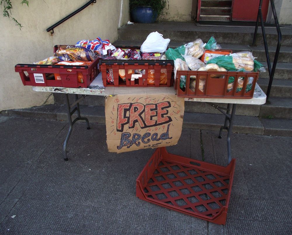 A colour photograph showing a table placed in front of steps at the entrance to a building. On the table are baskets filled with cellophane-packaged loaves and rolls. A white polythene bag, filled with unknown items, sits in one of the baskets. Attached to the table is a hand-written sign that reads ‘Free Bread’ and ‘Help yourselves!’. An empty basket lies on the ground in front of the table.