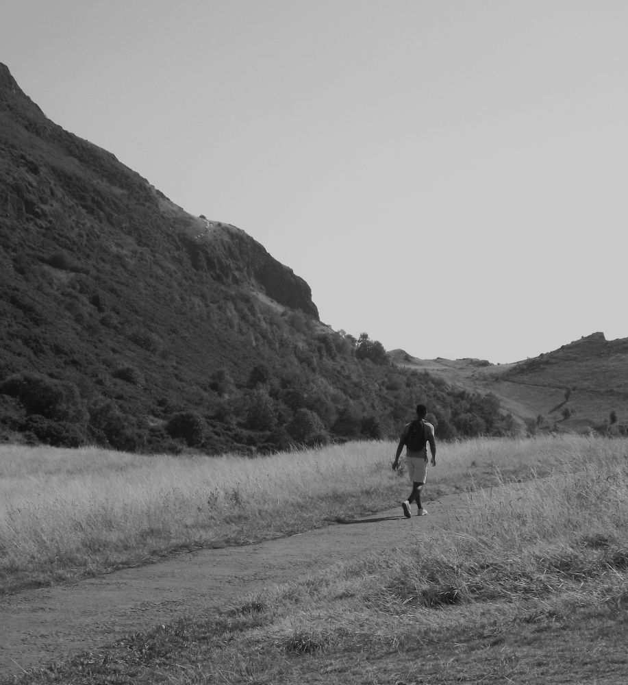 A monochrome photograph showing a young man in the middle distance. He is on a path that cuts through grass and is walking away from the camera. To his left and ahead of him are hills. The sky is cloudless.
