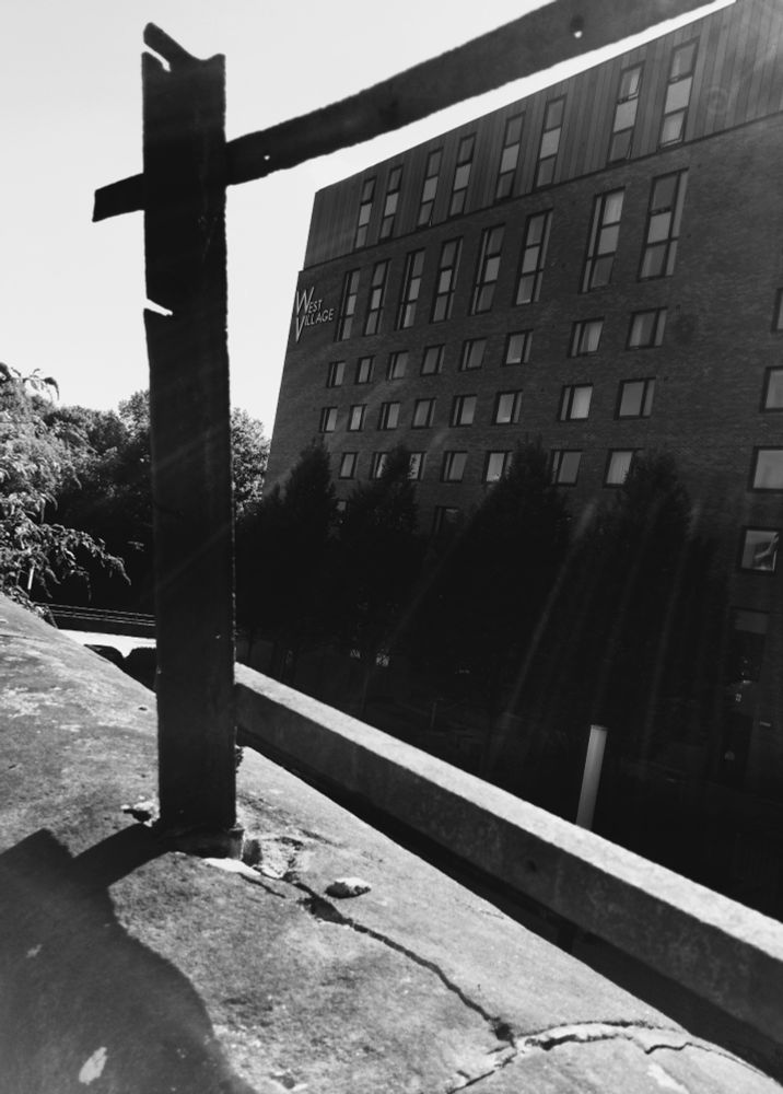 A monochrome photograph showing a modern student-accommodation building, ‘West Village’, viewed through part of a dilapidated fence.