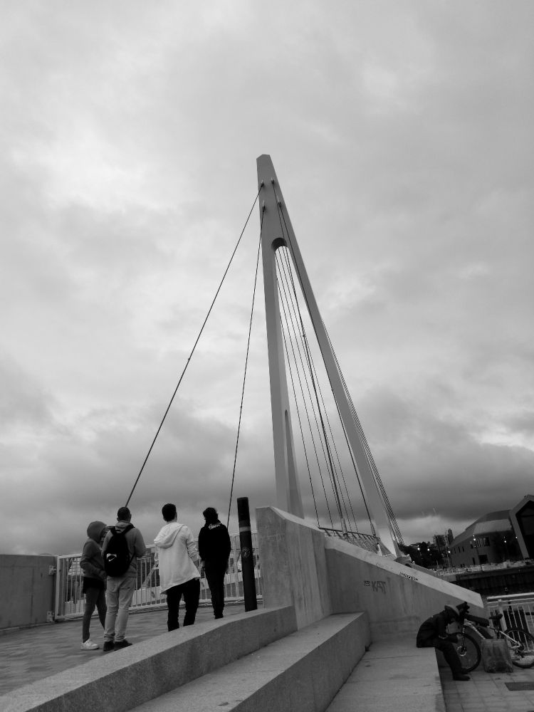 A monochrome photograph showing a small group of people waiting for a bridge over a river to re-open after it has swung shut. The sky is grey and clouded over. The scene has been photographed in such a way that everything, but especially the tower of the bridge, appears to be leaning to the left.