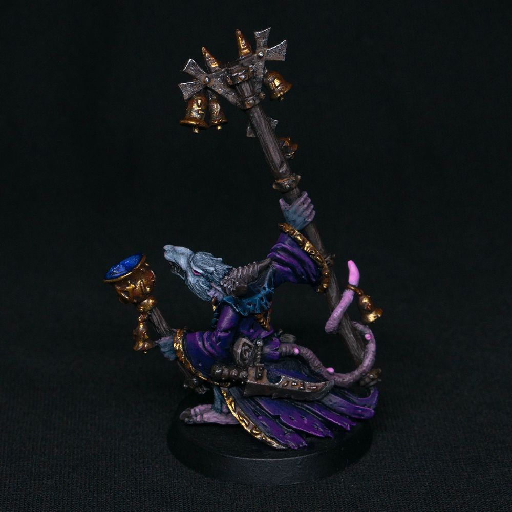 An older pewter Grey Seer miniature from Warhammer Fantasy Battles. This ambitious rat wizard has horns and light grey fur. He is adorned in purple robes with gold ornamentation and holds a golden bell in one hand and a staff in the other. Back view.