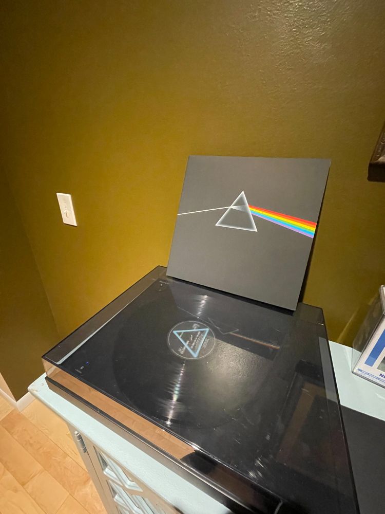 Image of a record player with a record and the sleeve of the record sitting on the record player leaning against the wall.  The record is Pink Floyd's "Dark Side of the Moon" showing a prism diffracting white light into a rainbow.