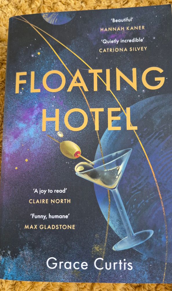 Image shows the cover of the book Floating Hotel by Grace Curtis, which has a picture of a cocktail glass, complete with an olive on a stck, floating in space