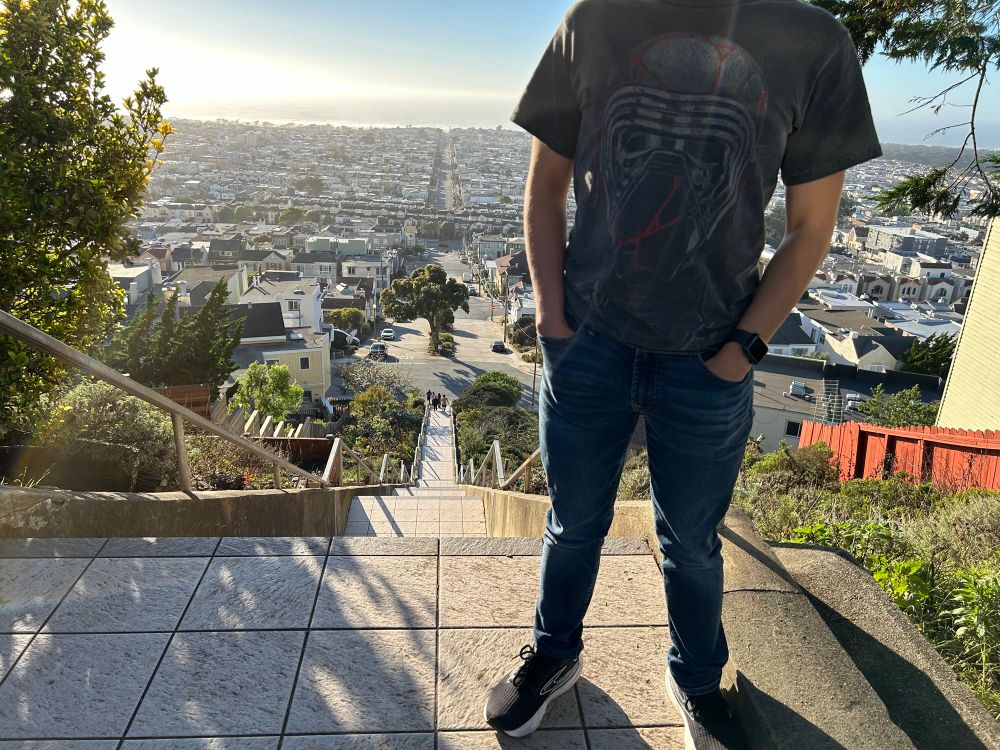 A standard t-shirt and jeans outfit with the shirt showcasing the visage of Kylo Ren/Ben Solo with his repaired helmet (red streaks included) from Star Wars: The Rise of Skywalker.

Behind is a large afternoon sunset vista of San Francisco (west side with Ocean Beach and the Pacific Ocean) at the top of the 16th Avenue Tiled Steps.
