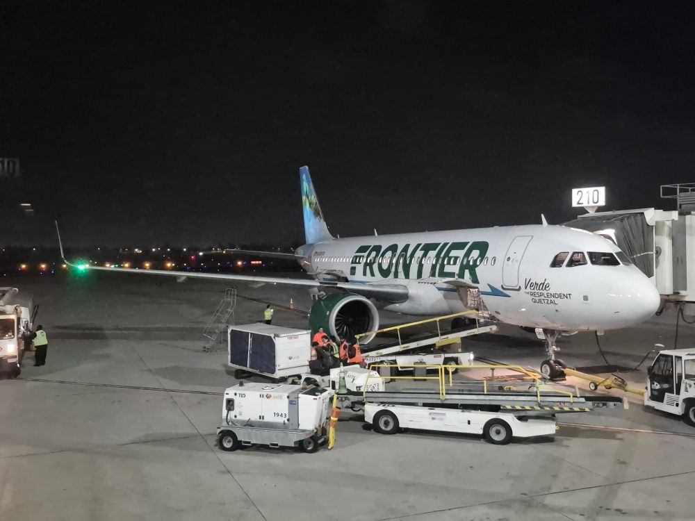 Photo of a Frontier Airlines Airbus A320neo at gate 210 at Ontario International Airport, at night. The text, "Verde the Resplendent Quetzal" is written under the cockpit windows, with a decal of the animal on the tailfin.