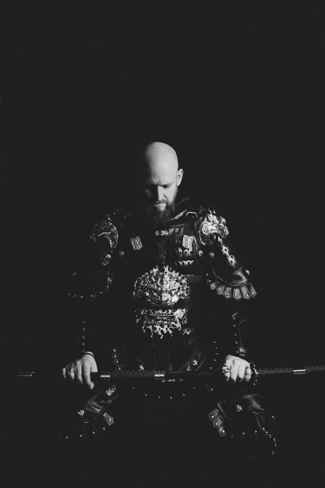 Studioaufnahme in schwarz/weiß, ein Mann sitzt in Samurai Rüstung mit einem Schwert in den Händen. Dunkler Hintergrund das Licht kommt von der rechten Seite.
