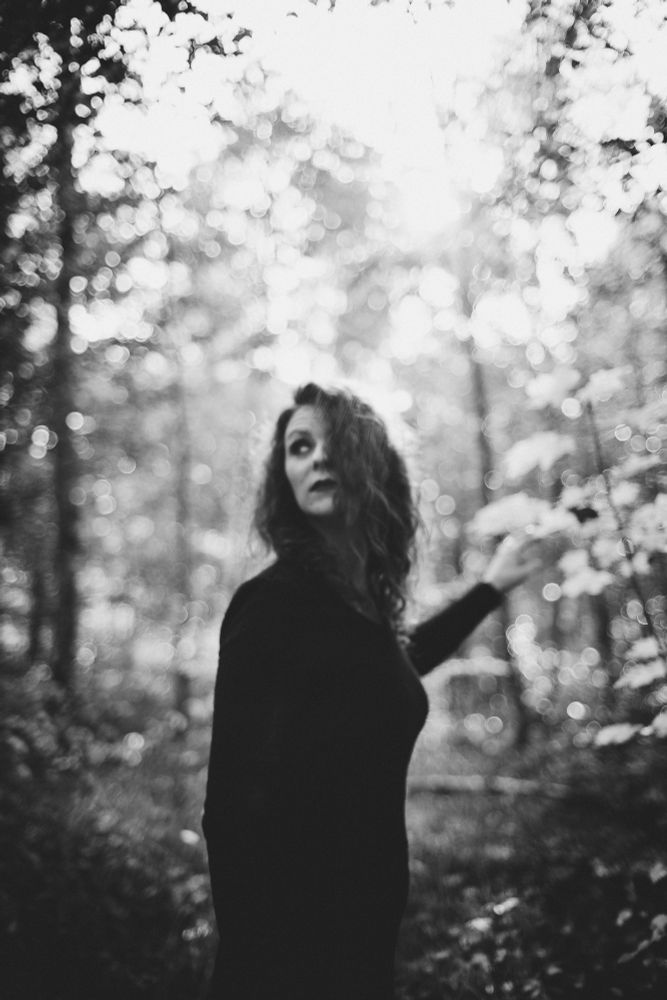Schwarz/Weiß Foto.
Eine Frau im schwarzen Kleid steht im Wald. Sie schaut schräg nach links oben. Und berührt mit einer Hand die Blätter hinter sich.
Das Foto ist gewollt etwas unscharf.