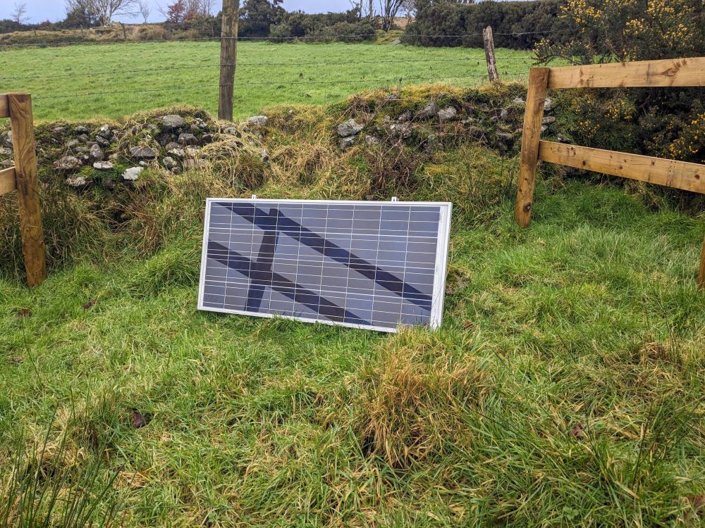 An image of a solar panel in a field in front of a stone wall. Grass has grown over the buried seismometer in the foreground